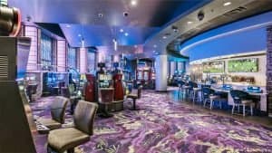 A modern casino floor featuring slot machines, ambient blue lighting, and a bar area with ceiling surveillance cameras