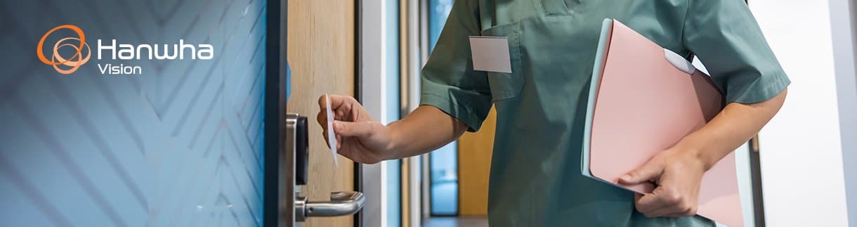 Healthcare employee using a key card with a commercial access control system at a secure office entrance while a security camera monitors entry.